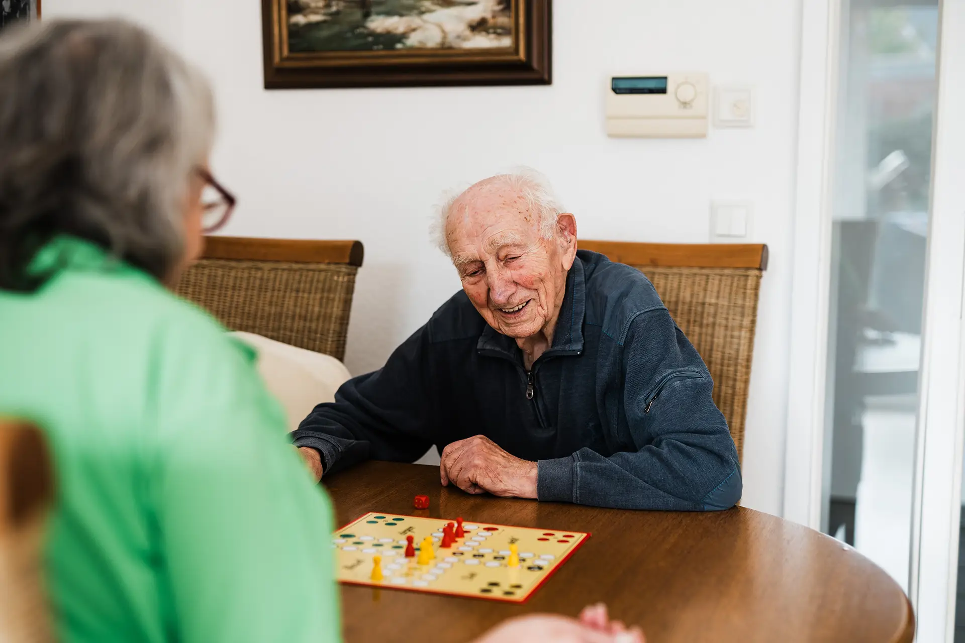 Senior spielt gemeinsam mit einer Betreuungsperson ein Gesellschaftsspiel im Rahmen der Alltagsbegleitung zu Hause