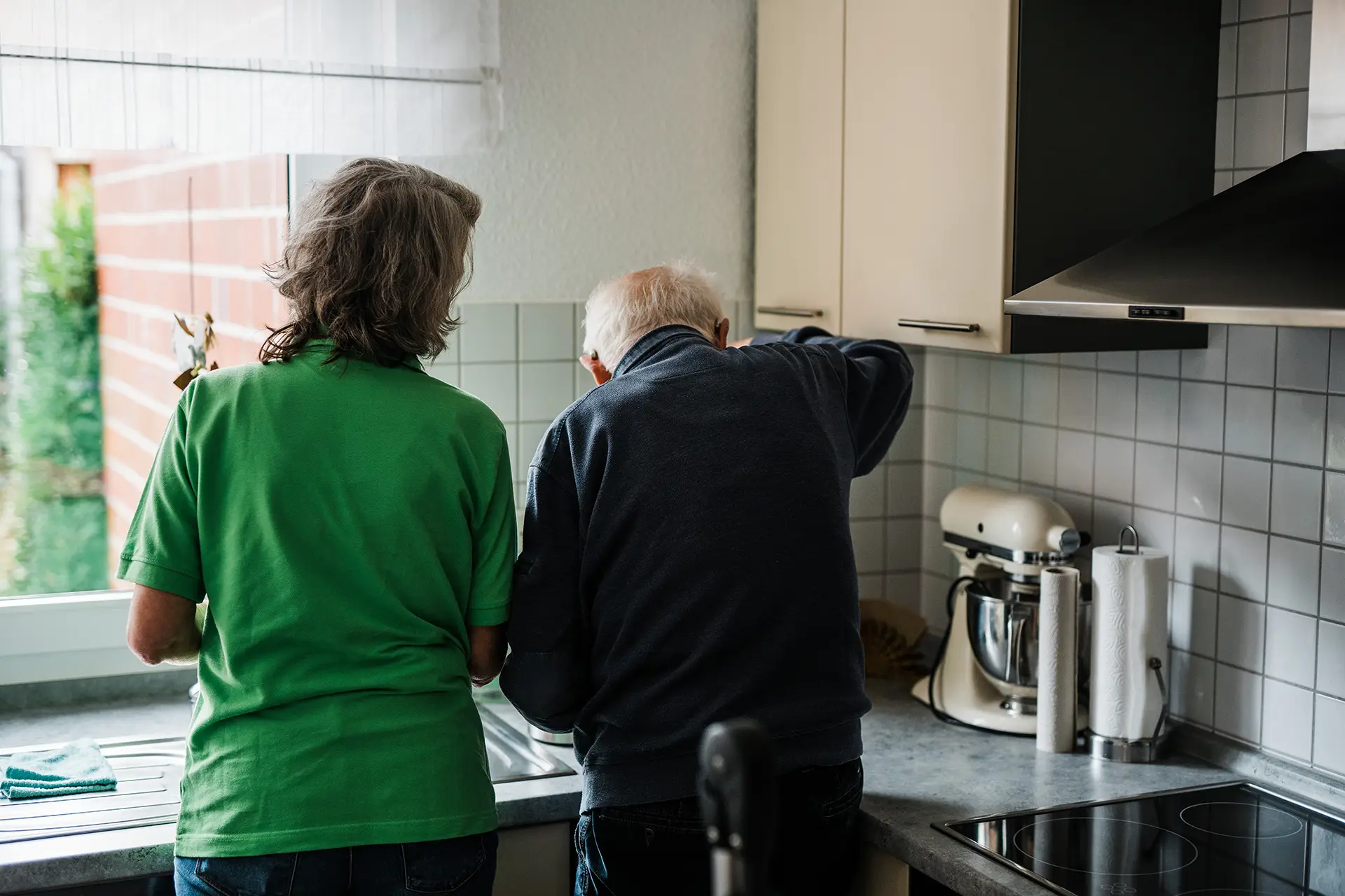 Betreuungsperson unterstützt einen Senior bei einer Tätigkeit in der Küche im Rahmen der Alltagshilfe zu Hause